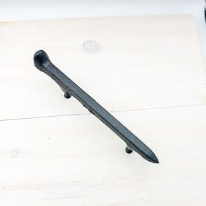 May include: A black, cast iron cabinet pull with a nail head design. The handle is a long, rectangular shape with a rounded head on one end and a pointed tip on the other. Two small screws are attached to the bottom of the handle.