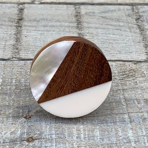 May include: A circular cabinet knob with a geometric design. It features dark wood, iridescent mother-of-pearl, and a white section. The knob is set against a grey wooden surface.