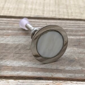 K&ouml;nnte beinhalten: Ein silberner Schrankknopf mit einer wei&szlig;en Perlmutteinlage. Der Knopf ist rund und hat eine glatte Oberfl&auml;che.