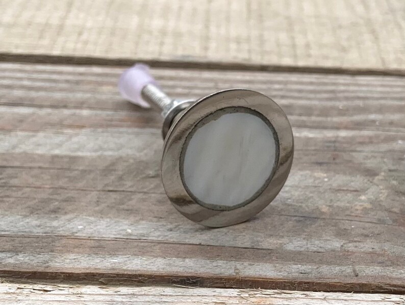 K&ouml;nnte beinhalten: Ein silberner Schrankknopf mit einer wei&szlig;en Perlmutteinlage. Der Knopf ist rund und hat eine glatte Oberfl&auml;che.