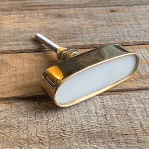 May include: An oval-shaped cabinet knob featuring a smooth white inlay surrounded by a polished gold-tone metal frame. The hardware includes a threaded metal bolt with a small nut and washer attached to the back for secure installation.