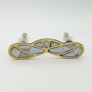 May include: A brass cabinet pull with a decorative design featuring inlaid mother-of-pearl pieces. The pull has a curved shape and two screw holes.
