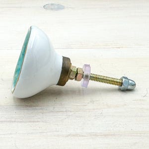 May include: A ceramic cabinet knob with a smooth white dome-shaped front and a teal-colored edge. The hardware includes a brass-toned metal base, a threaded bolt, a translucent plastic washer, and a silver-toned metal nut at the end of the bolt.