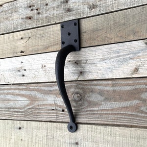 May include: Black, curved door pull with a square mounting plate. The handle is made of cast iron and is mounted on a rustic wooden surface. The door pull is designed for easy gripping and adds a touch of vintage charm.