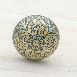 Peut inclure: Bouton de meuble rond et décoratif avec un motif floral et de feuilles complexe. Le bouton présente un fond bleu sarcelle avec un motif doré. Le motif symétrique recouvre toute la surface du bouton.