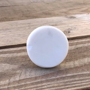 May include: A white, circular cabinet knob with a shiny surface. The knob is positioned centrally, set against a weathered, light brown wooden surface. The knob's smooth surface reflects the light.
