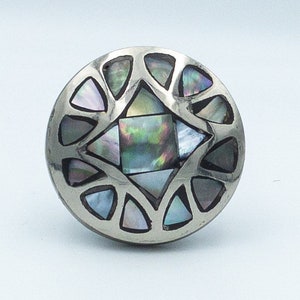 May include: A silver ring with a circular design featuring a central square of iridescent abalone shell surrounded by smaller triangular pieces of abalone shell.