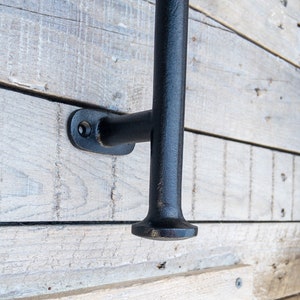 May include: Close-up of a black, cast iron door pull handle. The handle is mounted on a light-colored, weathered wooden surface. The handle has a T-shape design with a rounded base and a rectangular mounting plate.