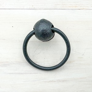 May include: A black, round, metal door knocker with a textured finish. The knocker features a circular handle attached to a decorative, spherical base. The hardware is designed for exterior doors and adds a rustic touch.