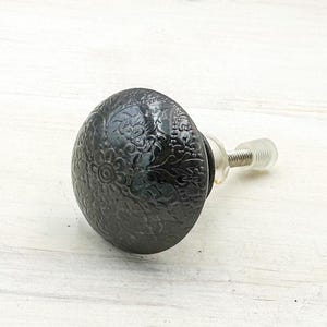 May include: A black, decorative cabinet knob with an embossed floral design. The knob has a rounded shape and a clear, threaded stem for attachment. The knob is made of a dark, possibly metal material.