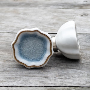 May include: Two white ceramic drawer pulls with a blue glaze and gold trim. The pull on the left has a scalloped edge and the pull on the right is round.
