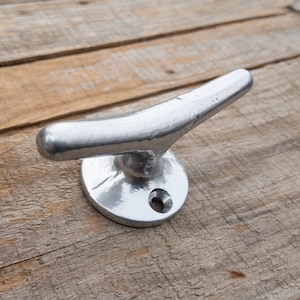 May include: A silver metal cleat hook with a rounded base. The hook is designed to be mounted on a wall or surface.