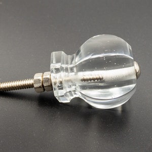 May include: A clear glass cabinet knob with a silver screw. The knob has a round, slightly textured shape.