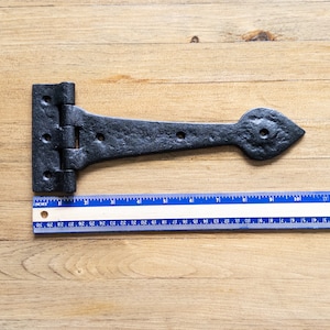 May include: Black metal hinge with a pointed end, measuring 7 inches in length. The hinge is shown on a wooden surface with a ruler for scale.
