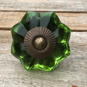 May include: A green glass cabinet knob with a flower-like design. The knob has a faceted, starburst shape with a bronze-coloured metal base. The knob is set against a weathered wood background.