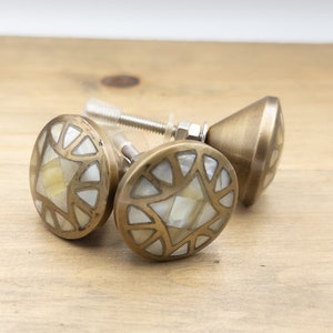 May include: Three brass cabinet knobs with mother-of-pearl inlay. The knobs have an oval shape with a geometric design and are mounted on screws. The knobs are set against a light brown wooden surface.