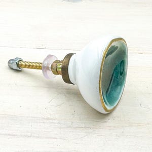 May include: A ceramic cabinet knob with a glossy white exterior and a deep teal interior basin. The knob features a gold-toned metal rim and a brass-colored mounting bolt with a clear plastic washer and a silver-toned metal nut at the end.