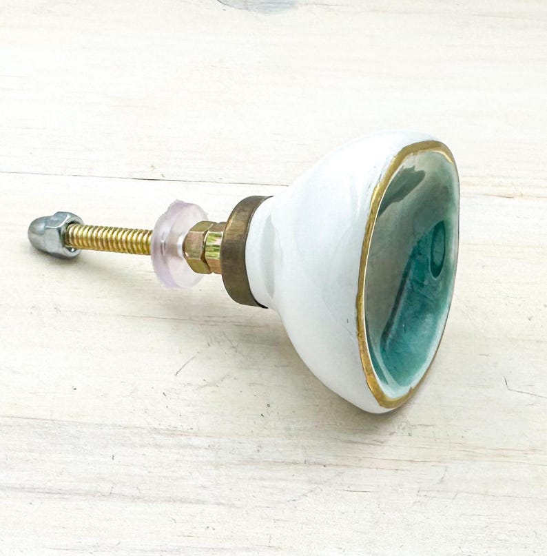 May include: A ceramic cabinet knob with a glossy white exterior and a deep teal interior basin. The knob features a gold-toned metal rim and a brass-colored mounting bolt with a clear plastic washer and a silver-toned metal nut at the end.