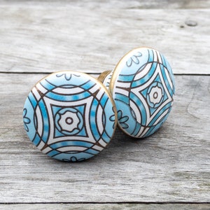 May include: Two ceramic cabinet knobs with a light blue and white geometric design. The knobs have a gold-coloured metal base and are set against a weathered wood background. The design features a central floral motif.