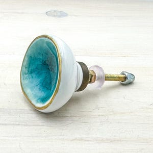 May include: A round ceramic cabinet knob featuring a concave center with a turquoise crackle glaze finish and a gold-toned rim. The exterior is smooth white ceramic, connected to a threaded metal bolt with a brass-colored base and a clear washer.