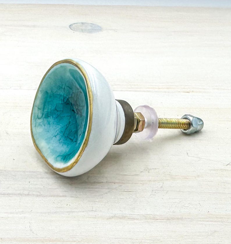 May include: A round ceramic cabinet knob featuring a concave center with a turquoise crackle glaze finish and a gold-toned rim. The exterior is smooth white ceramic, connected to a threaded metal bolt with a brass-colored base and a clear washer.