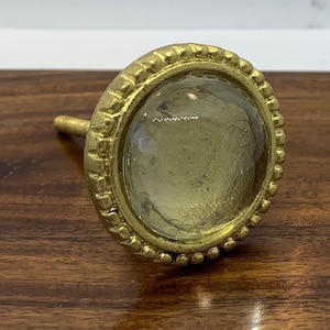 May include: A gold-toned cabinet knob with a clear glass centre. The knob has a decorative, beaded edge.
