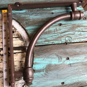 May include: A dark brown metal shelf bracket with a curved support arm and mounting plates featuring screw holes. A white measuring tape is positioned along the vertical side, indicating a height of approximately 26.7 cm for the bracket.