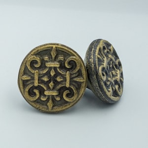 May include: Two ornate metal cabinet knobs with a dark brown finish and intricate designs. The knobs are square shaped with rounded edges and a raised centre.