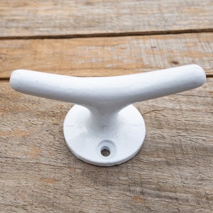 May include: A white cleat, a nautical hardware piece, is displayed against a wooden background. The cleat has a rounded base with a single hole and a curved top, measuring approximately 6 inches in length.