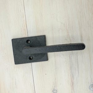 Peut inclure: Une poignée de porte noire avec une base carrée et une poignée rectangulaire texturée. La poignée comporte deux trous de vis et est faite d'un matériau sombre et mat. La poignée est posée sur une surface en bois de couleur claire.