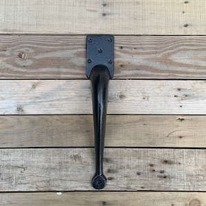 May include: Black metal table leg with a square top plate and a rounded bottom. The leg is attached to a wooden surface.