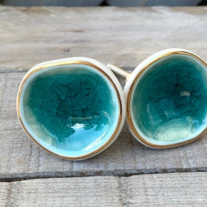 May include: Two round ceramic cabinet knobs with a concave, bowl-like shape. The interior surface features a teal crackle glaze finish, while the outer edges are trimmed with a thin gold metallic band. Each knob has a metal mounting bolt attached.