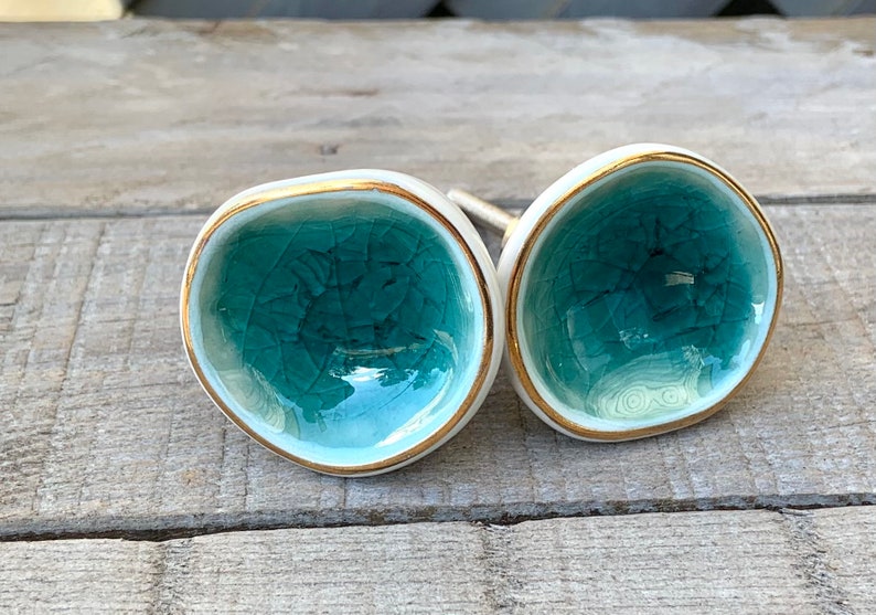 May include: Two round ceramic cabinet knobs with a concave, bowl-like shape. The interior surface features a teal crackle glaze finish, while the outer edges are trimmed with a thin gold metallic band. Each knob has a metal mounting bolt attached.