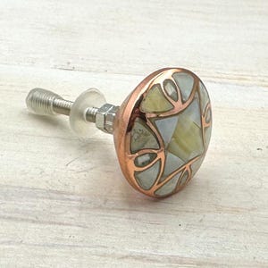 May include: A decorative cabinet knob with a copper-coloured rim and a mosaic design in shades of white, green, and yellow. The knob has a silver-coloured screw and nut for attachment. Suitable for drawers and cabinets.