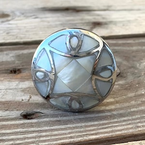 May include: A round, silver-toned cabinet knob with a white mother-of-pearl inlay. The inlay features a geometric design with a square in the center.
