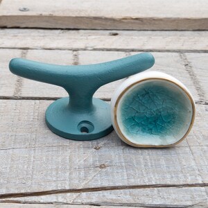 May include: A matte teal metal cleat hook with a circular base and a single screw hole, positioned next to a small, round, white ceramic knob featuring a crackled teal glaze interior and a thin gold-toned rim around the edge.