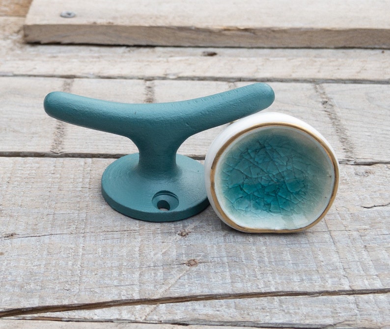 May include: A matte teal metal cleat hook with a circular base and a single screw hole, positioned next to a small, round, white ceramic knob featuring a crackled teal glaze interior and a thin gold-toned rim around the edge.