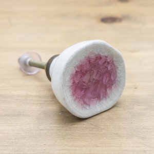 Peut inclure: Bouton de porte en céramique blanche avec un centre en verre rose et pailleté.