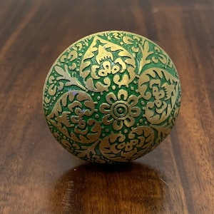 May include: A round, decorative cabinet knob with a green base and gold-colored floral and leaf pattern. The knob is made of metal and has a detailed, embossed design. The knob is a decorative hardware element.