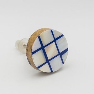 May include: A round, wooden cabinet knob with a blue and white checkered design. The knob has a white background with blue lines forming a grid pattern. The knob is made of wood and has a metal screw.