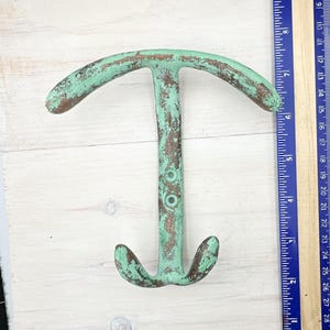 May include: A distressed, cast iron anchor-shaped wall hook with a weathered, turquoise finish. The hook has two curved arms at the top and a downward-facing hook at the bottom. Two holes are visible on the vertical center. The hook measures approximately 18 cm wide.