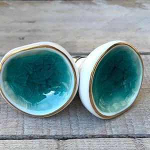 May include: Two ceramic cabinet knobs with a rounded, irregular shape. The center of each knob features a glossy, teal-colored crackle glaze finish, while the outer edges are smooth white ceramic trimmed with a thin, metallic gold-colored rim.