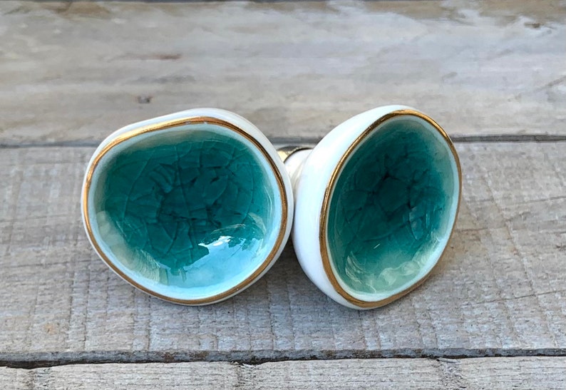 May include: Two ceramic cabinet knobs with a rounded, irregular shape. The center of each knob features a glossy, teal-colored crackle glaze finish, while the outer edges are smooth white ceramic trimmed with a thin, metallic gold-colored rim.