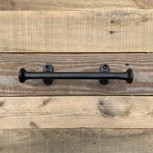 Peut inclure: Une barre à serviettes en fonte noire avec une finition texturée. La barre a des extrémités arrondies et est fixée sur une surface en bois. Le matériel de montage est visible. Le design est rustique et industriel.