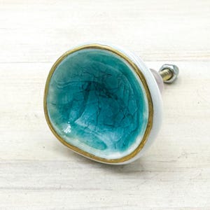 May include: A round ceramic cabinet knob featuring a concave center with a teal crackle glaze finish. The edge of the knob is trimmed with a metallic gold border, and the exterior side is white. A metal bolt with a nut extends from the back.
