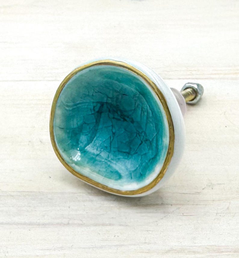 May include: A round ceramic cabinet knob featuring a concave center with a teal crackle glaze finish. The edge of the knob is trimmed with a metallic gold border, and the exterior side is white. A metal bolt with a nut extends from the back.