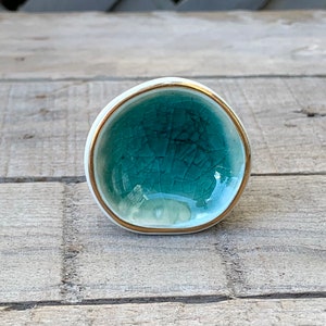 May include: A round ceramic knob with a teal crackle glaze finish on the front surface. The knob features a smooth white outer edge and a thin metallic gold rim bordering the teal center. The design is circular with a slightly irregular shape.