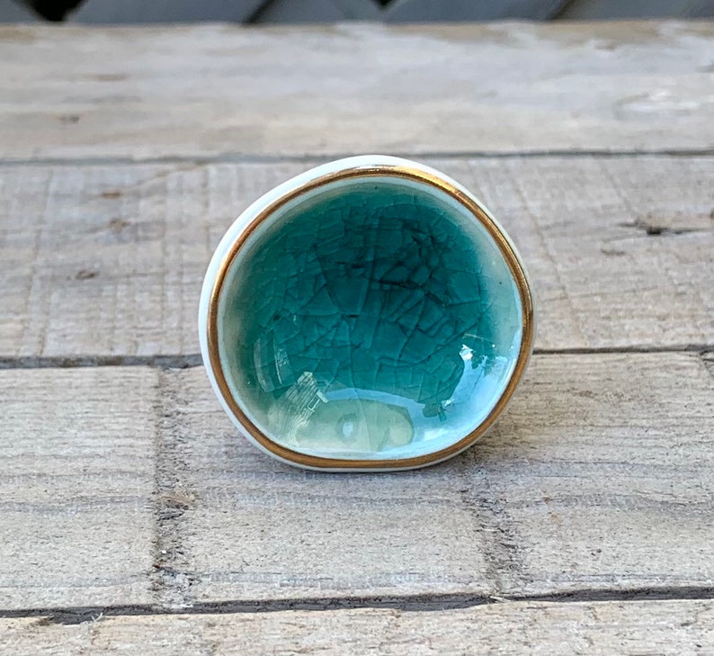 May include: A round ceramic knob with a teal crackle glaze finish on the front surface. The knob features a smooth white outer edge and a thin metallic gold rim bordering the teal center. The design is circular with a slightly irregular shape.