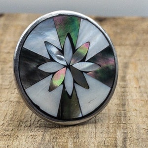 May include: A round, silver-toned ring with a black, white, and iridescent abalone shell inlay. The inlay features a stylised flower design.