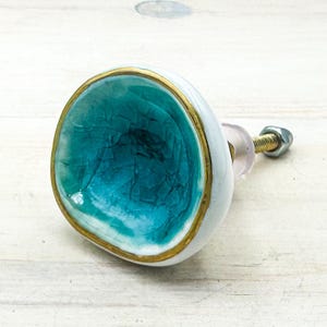 May include: A round cabinet knob with a concave center featuring a turquoise crackle glaze finish. The outer edge is painted with a metallic gold border, while the exterior side is white. A metal bolt and nut extend from the back of the knob.